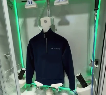 A navy blue pullover with "presso" logo is displayed on a mannequin inside a glass-enclosed, automated garment cleaning machine with green lighting at a pickup and delivery dry cleaning Katy location.