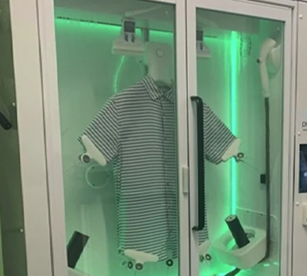 A striped short-sleeve shirt is being processed inside a glass-enclosed automated machine with green lighting and mechanical arms, showcasing innovative technology you'd expect from a top-rated laundry service near me.