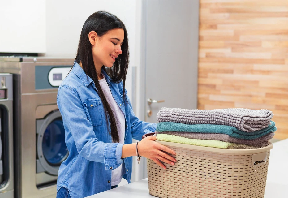 A woman places folded towels into a laundry basket in a laundromat.
