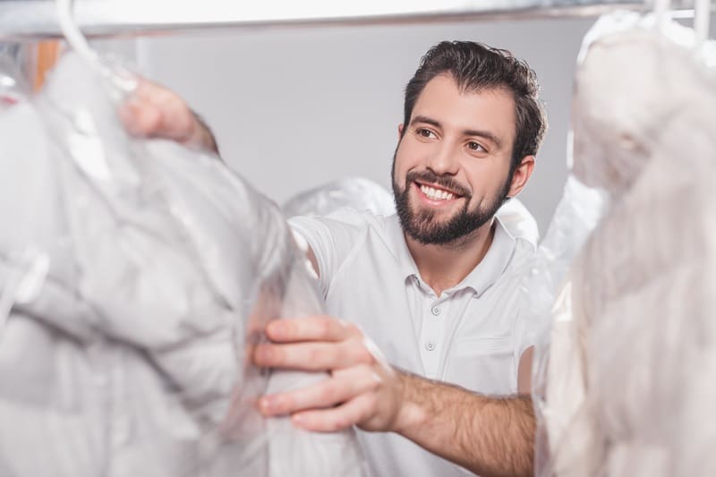 A man wearing a white polo shirt smiles while arranging clothes covered in plastic at a dry cleaning facility.
