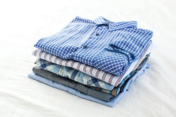 A neatly folded stack of shirts in different patterns and colors, including checks, stripes, and solids, placed on a white surface.