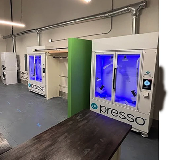 Two Presso machines with glass doors and blue interior lights stand side by side in a modern room, separated by a green divider. Pipes are visible on the wall behind them.