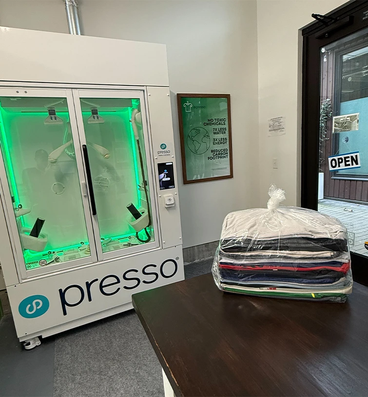A stack of folded clothes in a plastic bag sits on a counter next to a Presso laundry machine in a modern, minimalistic room.